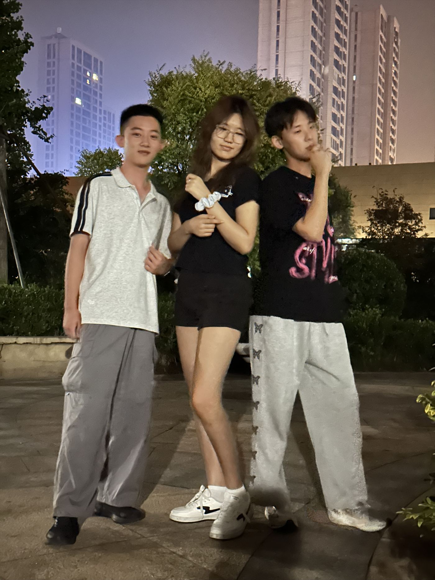 三个人站在夜晚的小区广场上合影，身后是亮着灯的高楼和树影